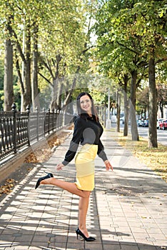 Portrait of a young elegant business woman indulging on the sidewalk among the trees in a black shirt and yellow skirt