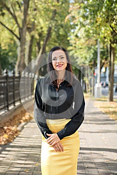 Portrait of a young elegant business woman standing on the sidewalk among the trees in a black shirt and yellow skirt