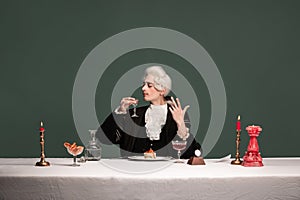 Portrait of young elegant man in peruke and vintage jacket sitting at table isolated on dark green background. Retro