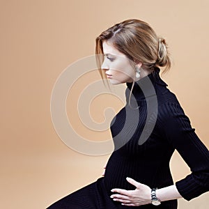 Portrait of young elegant pregnant woman in black clothes, sits on neutral background.