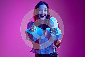 Portrait of young emotive girl holding two presents with happiness over pink background in neon light. Concept of