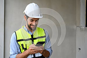 Portrait of young engineer in vest with white helmet standing on construction site, smiling and holding smartphone for worker,