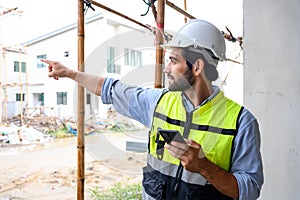 Portrait of young engineer in vest with white helmet standing on construction site, smiling and holding smartphone for worker,