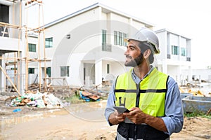Portrait of young engineer in vest with white helmet standing on construction site, smiling and holding smartphone for worker,