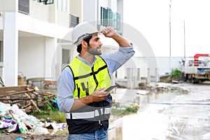 portrait of young engineer in vest with white helmet standing on construction site, smiling and holding smartphone for worker,