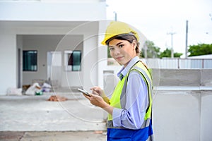 portrait of young engineer woman in vest with yellow helmet standing on construction site, smiling and holding smartphone for