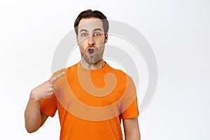 Portrait of young enthusiastic man looking surprised, pointing at himself, wearing orange t-shirt, white studio
