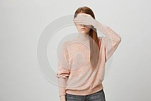 Portrait of young european ginger girl being calm and serious, covering eyes with hand, wearing trendy pullover and