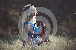 Portrait of a young fabulously beautiful girl in a dress with a violin sitting in dry grass on meadoe at the dawn, woman playing a