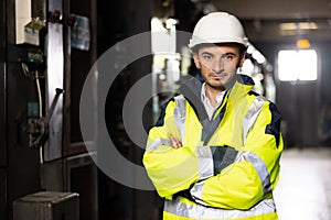 Portrait of young factory engineer or worker wearing safety vest and hard hat crossing arms at electrical control room