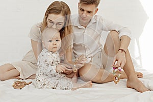 Portrait of young family with cherubic infant baby, with blue eyes holding wooden natural teether sitting on white bed. photo