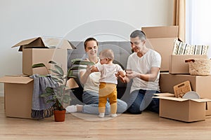 Portrait of young family unpacking moving box at their new apartment, sitting on floor near sofa and playing with their infant kid