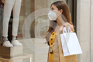 Portrait of young fashion woman with protective mask and shopping bags looking through shop window