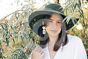 Portrait of young fashionable woman with modern earrings posing in  green olive branches in soft evening light, stylish boho girl