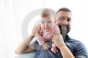 Portrait of young father with baby daughter sitting indoors on a sofa.