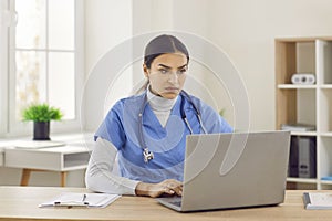Portrait of a young female doctor in blue medical uniform working in medical office on laptop.