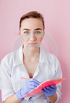 Portrait of young female doctor making notes in notebook on pink background. Medical industry concept