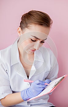 Portrait of young female doctor making notes in notebook on pink background. Medical industry concept