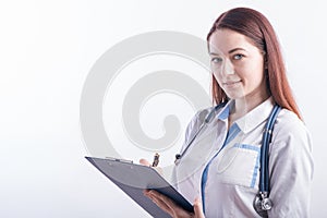 Portrait of a young female doctor in a white uniform with a folder and a pen in hands in the studio on a white background with cop