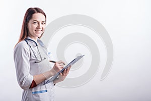 Portrait of a young female doctor in a white uniform with a folder and a pen in hands in the studio on a white background with cop