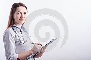 Portrait of a young female doctor in a white uniform with a folder and a pen in hands in the studio on a white background with cop