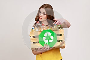 Portrait of young female holding box with plastic bottles and green recycling sing, pointing to