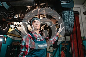 Portrait of a young female mechanic in uniform and gloves, posing near a car that is under repair. Bottom view. In the background