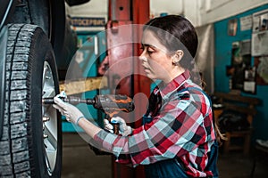 Portrait of a young female mechanic in uniform, tightening the nuts on the wheel disk. In the background there is an auto repair