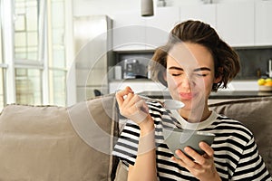 Portrait of young female model, student eating quick breakfast, holding bowl of granola or cereals with milk, sitting on