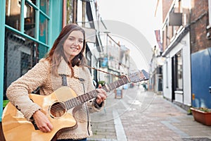 Portrait Of Young Female Musician Busking Playing Acoustic Guitar And Singing Outdoors In Street