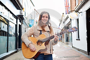 Portrait Of Young Female Musician Busking Playing Acoustic Guitar And Singing Outdoors In Street