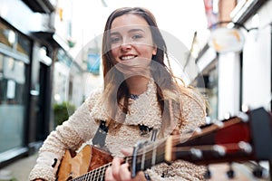 Portrait Of Young Female Musician Busking Playing Acoustic Guitar And Singing Outdoors In Street