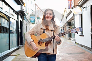 Portrait Of Young Female Musician Busking Playing Acoustic Guitar And Singing Outdoors In Street