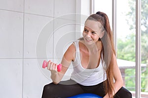 Portrait of young fitness sporty woman in sportswear sitting on fit ball and exercise with dumbbell in room. healthy lifestyle