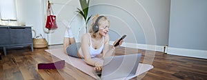 Portrait of young fitness woman, choosing workout on laptop, wearing headphones and using mobile phone exercise app