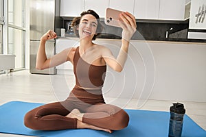 Portrait of young fitness woman, sitting at home on rubber mat, records video on smartphone, doing workout vlog, making