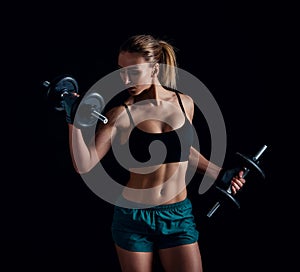 Portrait of a young fitness woman in sportswear doing workout with dumbbells on black background. Tanned athletic girl.