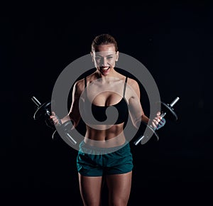 Portrait of a young fitness woman in sportswear doing workout with dumbbells on black background. Tanned athletic girl.