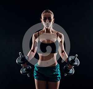 Portrait of a young fitness woman in sportswear doing workout with dumbbells on black background. Tanned athletic girl.