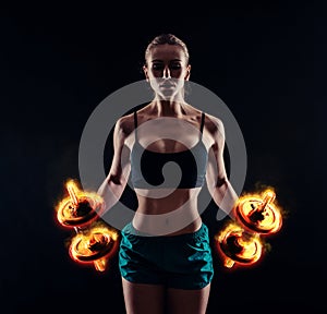 Portrait of a young fitness woman in sportswear doing workout with fiery dumbbells on black background. Tanned athletic girl.