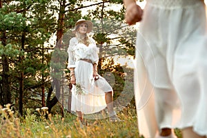 Portrait of young flawless woman standing in front of trees in forest in summer, watching little girl playing on grass.