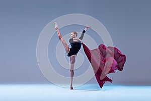 Portrait of young flexible ballerina dancing with deep red fabric isolated over blue grey studio background. Graceful