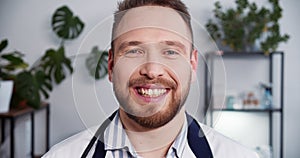 Portrait of young friendly Caucasian doctor man wearing white lab coat, stethoscope smiling at camera at light office.