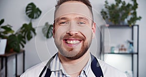 Portrait of young friendly Caucasian doctor man wearing white lab coat, stethoscope smiling at camera at light office.