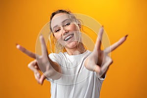 Portrait of a young funny woman showing peace gesture with two hands isolated over yellow background.