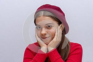 Portrait of a young girl in a beret and a red sweater, smiling mysteriously, propping her head with her hands, light background.