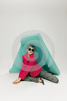 Portrait of young girl in bright jacket and vintage pants posing, sitting under big paper piece isolated over grey