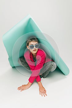 Portrait of young girl in bright jacket and vintage pants posing, sitting under big paper piece isolated over grey