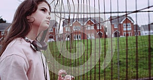 Portrait of young girl dressed in pink vest near rusty metallic fence. Girl have headphones and thinking while staring