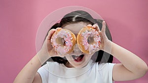 Portrait young girl makes glasses from sweet donuts with icing in her hands, smiling, looks at camera. Pink background
